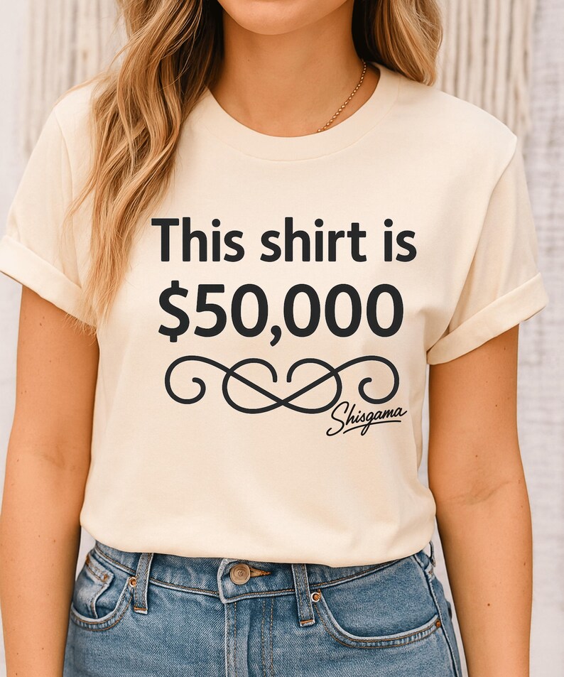 May include: Cream-colored t-shirt with the text "This shirt is $50,000" in black, along with a decorative swirl and the word "Shisgama" in script. The shirt has rolled-up sleeves and is worn with blue jeans.
