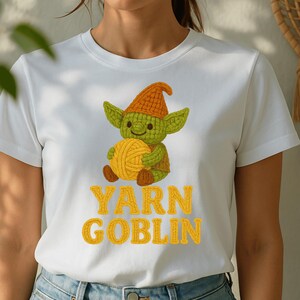 May include: White t-shirt featuring a green goblin character holding a ball of yellow yarn. The goblin wears an orange hat. The words "YARN GOBLIN" are printed in yellow block letters below the goblin illustration.
