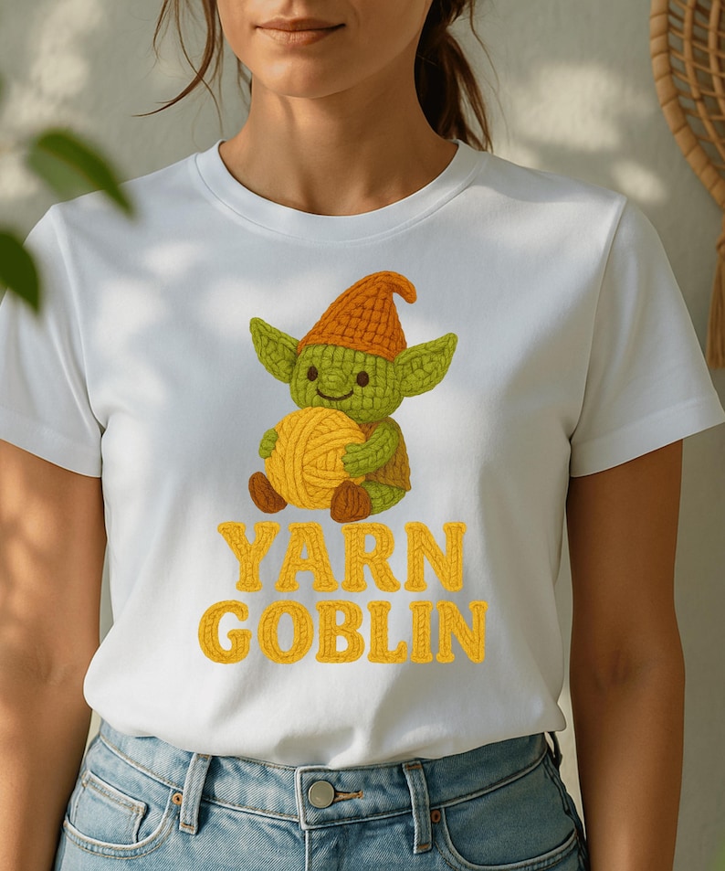 May include: White t-shirt featuring a green goblin character holding a ball of yellow yarn. The goblin wears an orange hat. The words "YARN GOBLIN" are printed in yellow block letters below the goblin illustration.