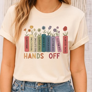 Può includere: T-shirt color crema con grafica di libri e fiori colorati. I libri presentano testi come "IMMIGRANTS" e "HANDS OFF". La maglietta è realizzata in materiale morbido.