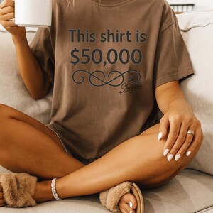 May include: Brown t-shirt with the text "This shirt is $50,000" in dark gray. The shirt has a decorative infinity symbol and the word "Shigama" below the text. The person is holding a white mug.
