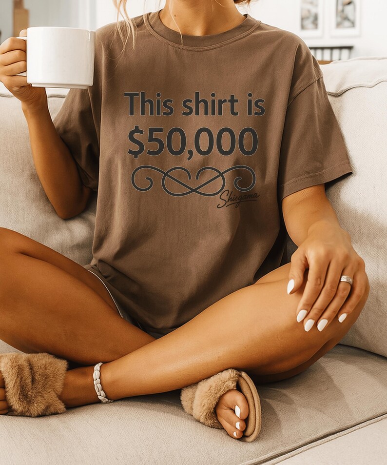May include: Brown t-shirt with the text "This shirt is $50,000" in dark gray. The shirt has a decorative infinity symbol and the word "Shigama" below the text. The person is holding a white mug.