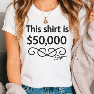 May include: White t-shirt with the text "This shirt is $50,000" in black, along with a decorative swirl and the word "Shisgama" in a script font. The shirt is short-sleeved.