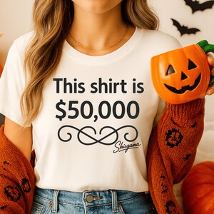 May include: White t-shirt with the text "This shirt is $50,000" and a decorative swirl design, with the brand name "Shisgama" below. The person is wearing blue jeans and an orange sweater. A ceramic pumpkin mug is held in the right hand.