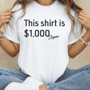 Può includere: T-shirt bianco con la scritta "This shirt is $1,000 Shisgama" in nero. La maglietta è indossata con jeans blu e scarpe da ginnastica bianche. La modella indossa una collana e braccialetti dorati.