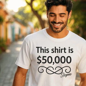 May include: White t-shirt with the text "This shirt is $50,000" in black. The shirt also features a decorative swirl design and the word "Shisgama" in script. The shirt is being worn by a person.
