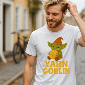 May include: White t-shirt featuring a cartoon goblin holding a ball of yarn. The goblin wears a green face and a brown hat. The text "YARN GOBLIN" is printed in yellow below the graphic.