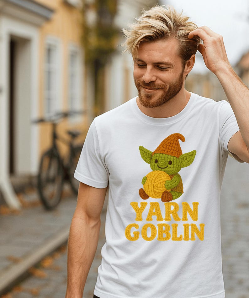 May include: White t-shirt featuring a cartoon goblin holding a ball of yarn. The goblin wears a green face and a brown hat. The text "YARN GOBLIN" is printed in yellow below the graphic.