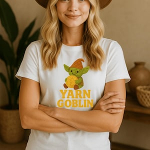 May include: White t-shirt featuring a cartoon goblin holding a ball of yarn. The goblin is green with a brown hat. The text "YARN GOBLIN" is in yellow. The shirt is short-sleeved.