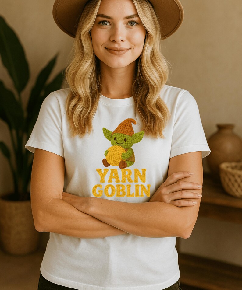 May include: White t-shirt featuring a cartoon goblin holding a ball of yarn. The goblin is green with a brown hat. The text "YARN GOBLIN" is in yellow. The shirt is short-sleeved.