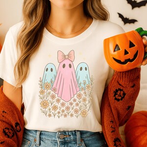 Può includere: T-shirt color crema con un disegno di tre fantasmi pastello e fiori. Il fantasma centrale ha un fiocco rosa. La persona tiene in mano una tazza arancione a forma di zucca. I pipistrelli sono sullo sfondo.