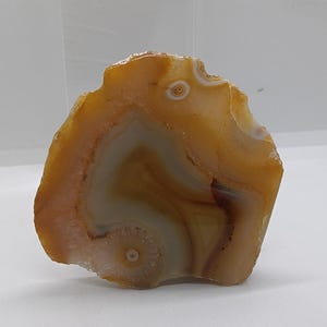 Può includere: Una fetta di pietra d'agata lucidata con una forma naturale e irregolare. La pietra presenta bande di colori arancione, marrone e crema, con un aspetto traslucido. L'agata ha una superficie liscia e lucida, probabilmente per scopi decorativi.
