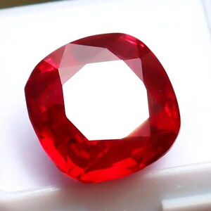 May include: A vibrant, cushion-cut red gemstone with a faceted surface that reflects light, creating a bright white centre. The gemstone is securely positioned within a small, square, white plastic display box with a smooth, matte finish.