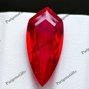 Könnte beinhalten: Ein leuchtend roter Edelstein mit einer schildartigen, länglichen Fünfeckform und zahlreichen Facetten, die das Licht reflektieren. Der Stein hat eine spitz zulaufende Unterseite und eine flache, abgewinkelte Oberkante in einem tiefen Karmesinrot.