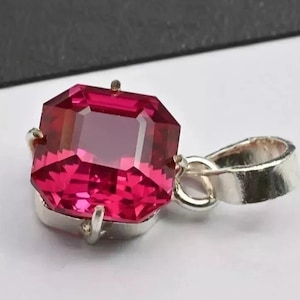 May include: A square-cut, deep pink gemstone pendant set in a polished silver-toned metal frame. The pendant features a faceted stone with sharp corners and a prominent bail attached to the side, designed for suspension on a chain or cord.