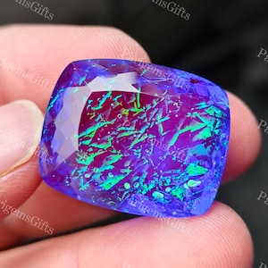 May include: A square-cut gemstone with a vibrant display of colors. The gem features a deep purple base with flashes of green and blue, creating an iridescent effect. The facets catch the light, enhancing the stone's brilliance. The gem is held between fingers.