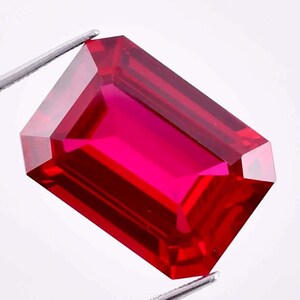 May include: A vibrant, deep red gemstone cut into a rectangular emerald shape with sharp, clean facets. The stone is held by silver-toned metal tweezers, showcasing its translucent clarity and intense, saturated crimson colour throughout the piece.