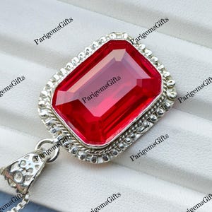 May include: A rectangular pendant featuring a large, faceted, vibrant red gemstone set in a silver-toned metal frame with a twisted rope border. The pendant includes a decorative, perforated bail and is attached to a thin, silver-toned chain.