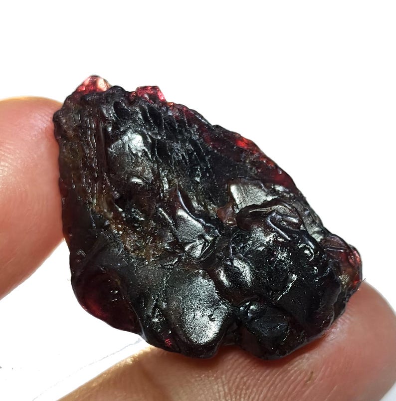 May include: A rough, dark red gemstone with a glossy, uneven surface. The natural mineral has a deep, almost black color in some areas, with hints of brighter red at the edges. The stone is held between fingers, showcasing its raw, unpolished form.
