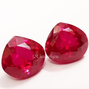May include: Two faceted, heart-shaped, deep red gemstones. The gems are highly polished, reflecting light, and are set against a white background. The gems are likely rubies, and the image is well-lit, showcasing their color and clarity.