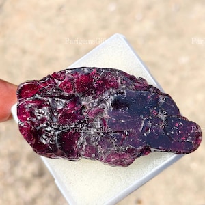 Può includere: Una gemma grezza, bordeaux scuro, con una superficie strutturata. La pietra è tenuta in un contenitore quadrato bianco. La gemma ha un aspetto scuro, quasi nero, in alcune aree.