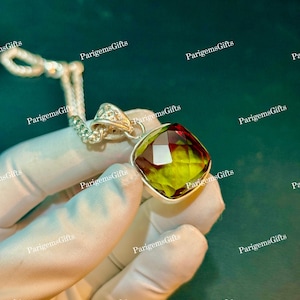 May include: A pendant necklace featuring a square-cut, faceted gemstone with a gradient of yellow, green, and deep red tones. The stone is set in a silver-toned metal frame with a decorative, filigree-style bail attached to a silver-toned chain.