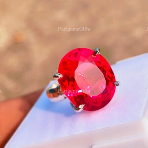 May include: A silver ring with a large, oval-cut, bright red gemstone. The gemstone is the focal point, with a faceted surface that catches the light. The ring is set on a white surface, with the text "ParigemsGifts" visible.