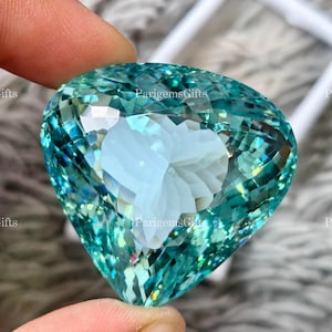 May include: A large, heart-shaped gemstone with a faceted surface. The gem is a vibrant teal colour with a lighter, almost white, heart-shaped area in the centre. The text "ParigemsGifts" is visible on the surface.
