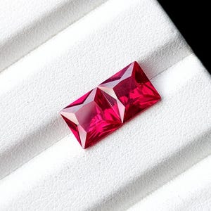 May include: Two vibrant, deep red gemstones with a rectangular princess cut. Each stone features sharp, geometric facets that reflect light, creating a brilliant, saturated appearance with clear, defined edges and a polished, smooth surface finish.