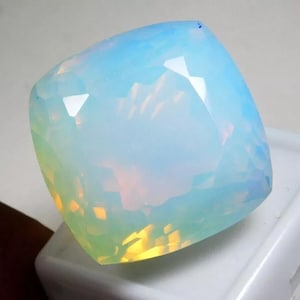May include: A cushion-cut gemstone with a translucent, milky appearance. The stone displays a soft, pale blue base color with internal flashes of yellow and orange light reflecting through its faceted surface, creating a shimmering effect.