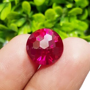 May include: A faceted, round, ruby-red gemstone with the text "VictoriaFineGem" visible. The gem is held between fingers, with a blurred green background.