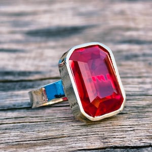 以下が含まれることがあります： 大きな長方形の赤い宝石が付いたシルバーリング。リングは青いアクセントが付いたシルバーバンドです。宝石が焦点となり、光を捉え、周囲の環境を反射しています。