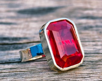 Peut inclure: Une bague en argent avec une grosse pierre rouge rectangulaire à facettes. La bague a un anneau argenté avec un accent bleu. La pierre précieuse est le point central, captant la lumière et reflétant l'environnement.