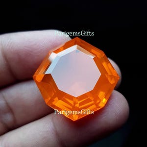 May include: A faceted, octagonal orange gemstone held in a hand. The gem has a clear, reflective surface and is labelled "ParigemsGifts". The gem's facets catch the light, creating a brilliant display. The gemstone is a vibrant, eye-catching piece.