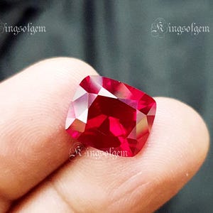 Könnte beinhalten: Nahaufnahme eines facettierten Rubins im Kissenschliff. Der Edelstein ist ein tiefes, leuchtendes Rot mit vielen Facetten, die das Licht reflektieren. Der Text "Ringsofgem" ist oben links und rechts zu sehen.