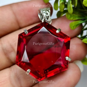May include: A large, faceted, hexagonal red gemstone pendant with a silver bail. The gemstone is a deep, vibrant red color and is set in a silver metal setting. The pendant is a striking piece of jewelry.