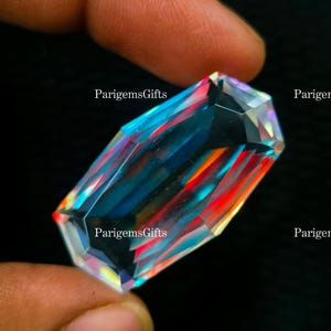 May include: A large, rectangular gemstone with a multifaceted cut. The gem displays a spectrum of colors, including blue, red, and yellow, with the text "ParigemsGifts" visible on the surface. The gem is held between fingers against a dark background.