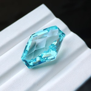 May include: A faceted, kite-shaped gemstone in a vibrant, translucent light blue colour. The stone features a geometric cut with multiple triangular and trapezoidal facets that reflect light, creating a clear, brilliant appearance throughout.