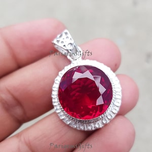 Op de afbeelding: Een zilveren hanger met een grote, gefacetteerde, dieprode edelsteen. De ronde edelsteen is gezet in een decoratieve zilveren zetting. De hanger heeft een getextureerde beugel voor een ketting. Elegant ontwerp.