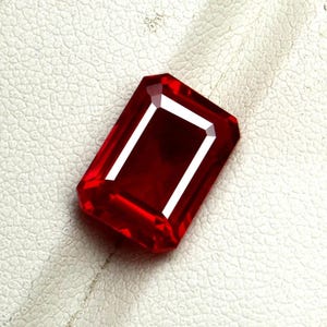 Può includere: Una gemma di un rosso profondo e vibrante, tagliata in una forma rettangolare a smeraldo. La pietra presenta sfaccettature nette e pulite che riflettono la luce, creando riflessi bianchi brillanti lungo i bordi e un ricco colore cremisi.