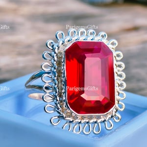 May include: A silver ring featuring a large, rectangular, vibrant red gemstone. The gemstone is set in a decorative, ornate silver frame with a scalloped edge. The ring is displayed on a light blue surface.