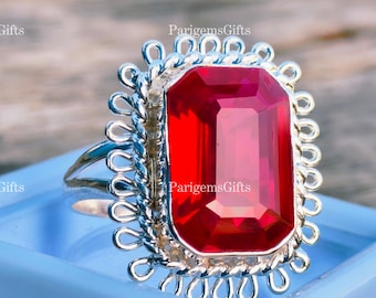 Peut inclure: Une bague en argent ornée d'une grande pierre rouge vif de forme rectangulaire. La pierre est sertie dans un cadre argenté orné avec un bord festonné. La bague est présentée sur une surface bleu clair.