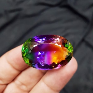 Op de afbeelding: Een ovaalvormige edelsteen met een veelkleurig kleurenpalet, variërend van groen tot paars en oranje. Het gefacetteerde oppervlak reflecteert het licht, wat een schitterend effect creëert. De edelsteen wordt in een hand gehouden.
