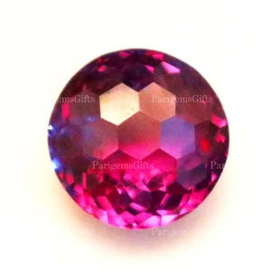 May include: A faceted, round gemstone with a vibrant color gradient. The gem transitions from deep magenta to a lighter pink and purple hue. The surface reflects light, showcasing the intricate cuts. The text "ParigemsGifts" is visible on the gem.