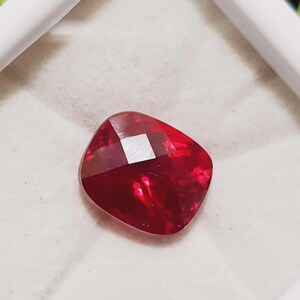 Peut inclure: Une pierre précieuse rubis taillée en coussin. La gemme est d'un rouge profond avec une facette carrée et réfléchissante au centre. La pierre est posée sur un fond blanc dans un cadre blanc.