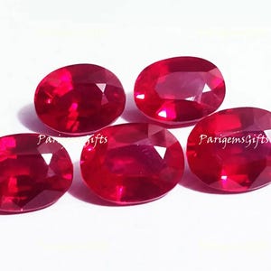 May include: Five oval-cut, faceted rubies in varying sizes are displayed against a white background. The gemstones are a deep, vibrant red, with visible internal reflections. The text "ParigemsGifts" is visible on some of the gems.