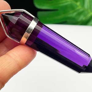 May include: A double-terminated, deep purple amethyst crystal pendant featuring a smooth, faceted hexagonal shape. The crystal is encased in a polished silver-toned metal cap with a small circular bail at the top for hanging as a necklace.