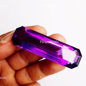Könnte beinhalten: Ein facettierter, rechteckiger Amethyst-Edelstein mit tiefvioletter Farbe. Der Edelstein wird in einer Hand gehalten, wodurch seine polierten Oberflächen und seine Klarheit zur Geltung kommen. Der Text "ParigemsGifts" ist sichtbar.