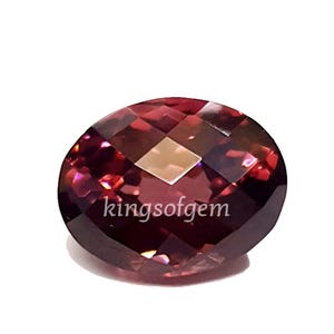 Può includere: Una gemma sfaccettata, di forma ovale e di colore bordeaux intenso. La superficie lucida presenta molteplici sfaccettature che riflettono la luce. Il testo "kingsof gem" è visibile sulla superficie.