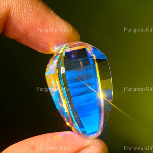May include: A faceted, teardrop-shaped gemstone with a vibrant blue interior and clear, reflective exterior. The gem is held between two fingers, showcasing its intricate cuts and the play of light across its surface. The background is a blurred green.